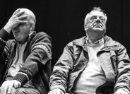 Black and white image of 2 men sitting beside each other the man on the left has his face covered with his hand and the man on the right is leaning his head back with closed eyes