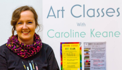 Image for art classes featuring the facilitator and artist Caroline Keane smiling beside her art class posters