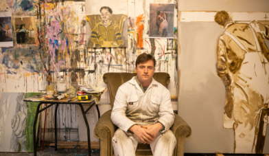 P Addy Critchley sitting in his painting studio in white overalls with large paintings behind him