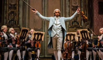 Chevalier movie still of violinist standing with arms outstretched in a room of seated violin players