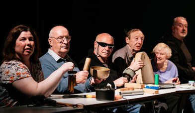6 people sit in a row in a dark room at a table One holds a bodhran and one has dark glasses on
