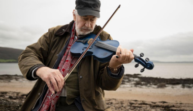 An older man plays the fiddle on a beach as the image for the irish film fidil ghorm