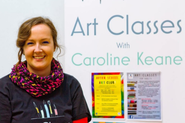 Image for art classes featuring the facilitator and artist Caroline Keane smiling beside her art class posters