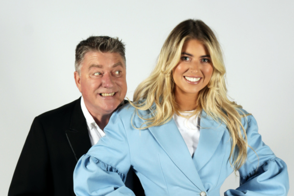 Image features Pat Shortt smiling at Faye Shortt who is dressed in a blue suit and they are standing in front of a bright white background