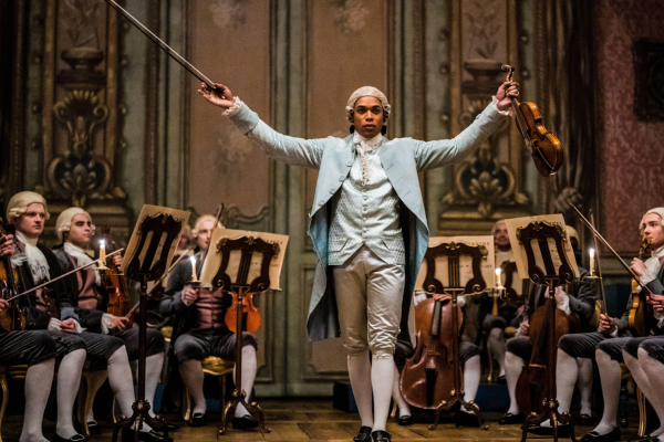 Chevalier movie still of violinist standing with arms outstretched in a room of seated violin players