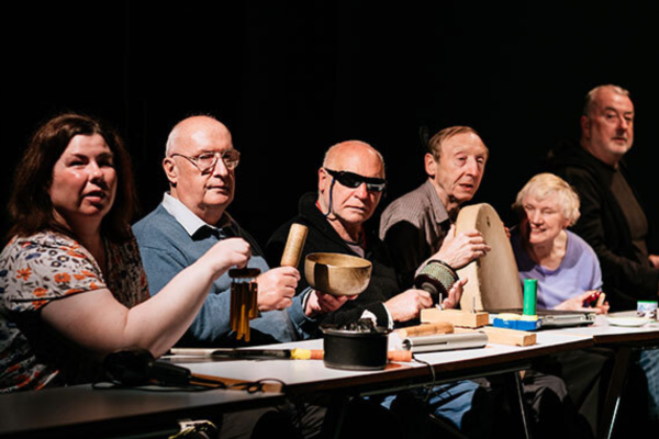 6 people sit in a row in a dark room at a table One holds a bodhran and one has dark glasses on