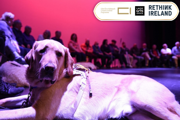 Golden lab guide dog sits on a stage with people in the distant background